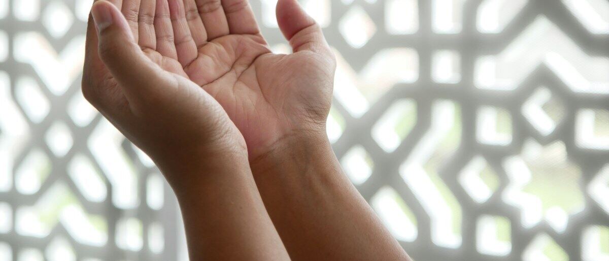 Duas mãos abertas erguidas em súplica diante de um painel rendado de luz