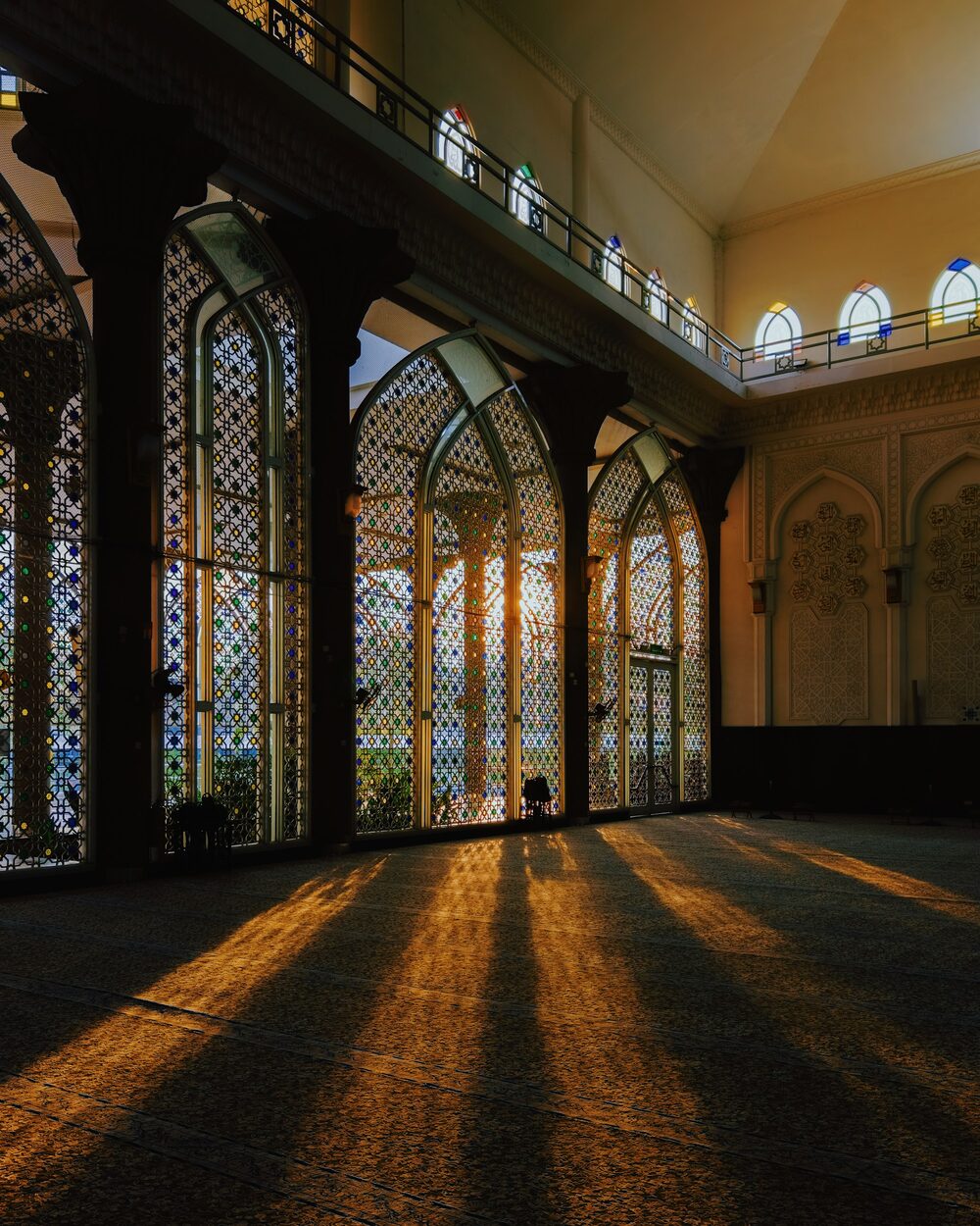 Luz dorada del atardecer que entra por altas ventanas en arco sobre la alfombra de la sala de oración de una mezquita