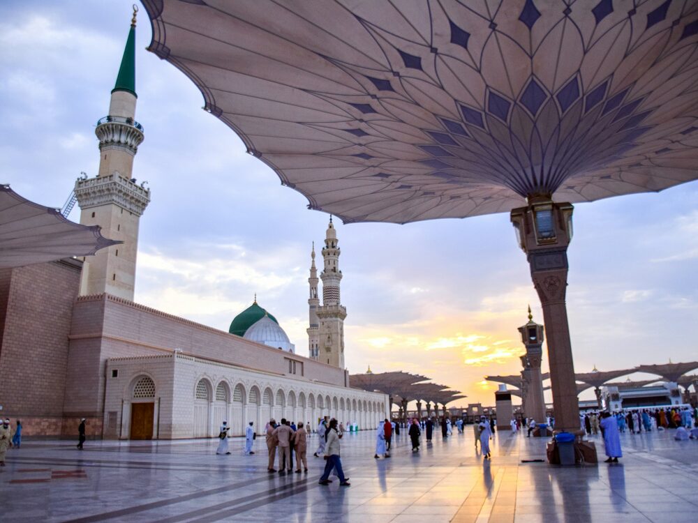 Personas que caminan bajo las grandes sombrillas del patio de la Mezquita del Profeta en Medina al atardecer